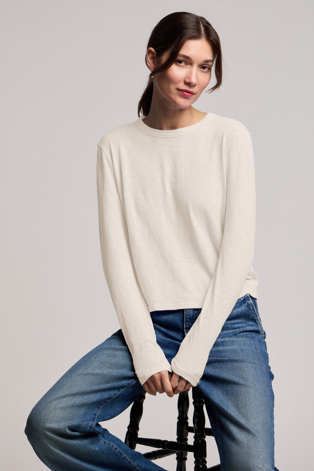 A woman wearing the MEGAN TEE by Velvet by Graham & Spencer and blue jeans sits on a stool against a plain background.-40808429420737