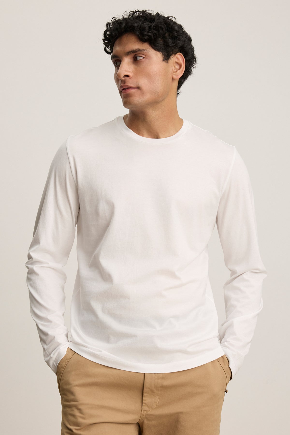 A man wears the ALFIE TEE by Velvet by Graham & Spencer—a midweight mercerized cotton long sleeve—in tan pants, standing against a neutral background with his hands in his pockets, looking to the side.-39450513047745