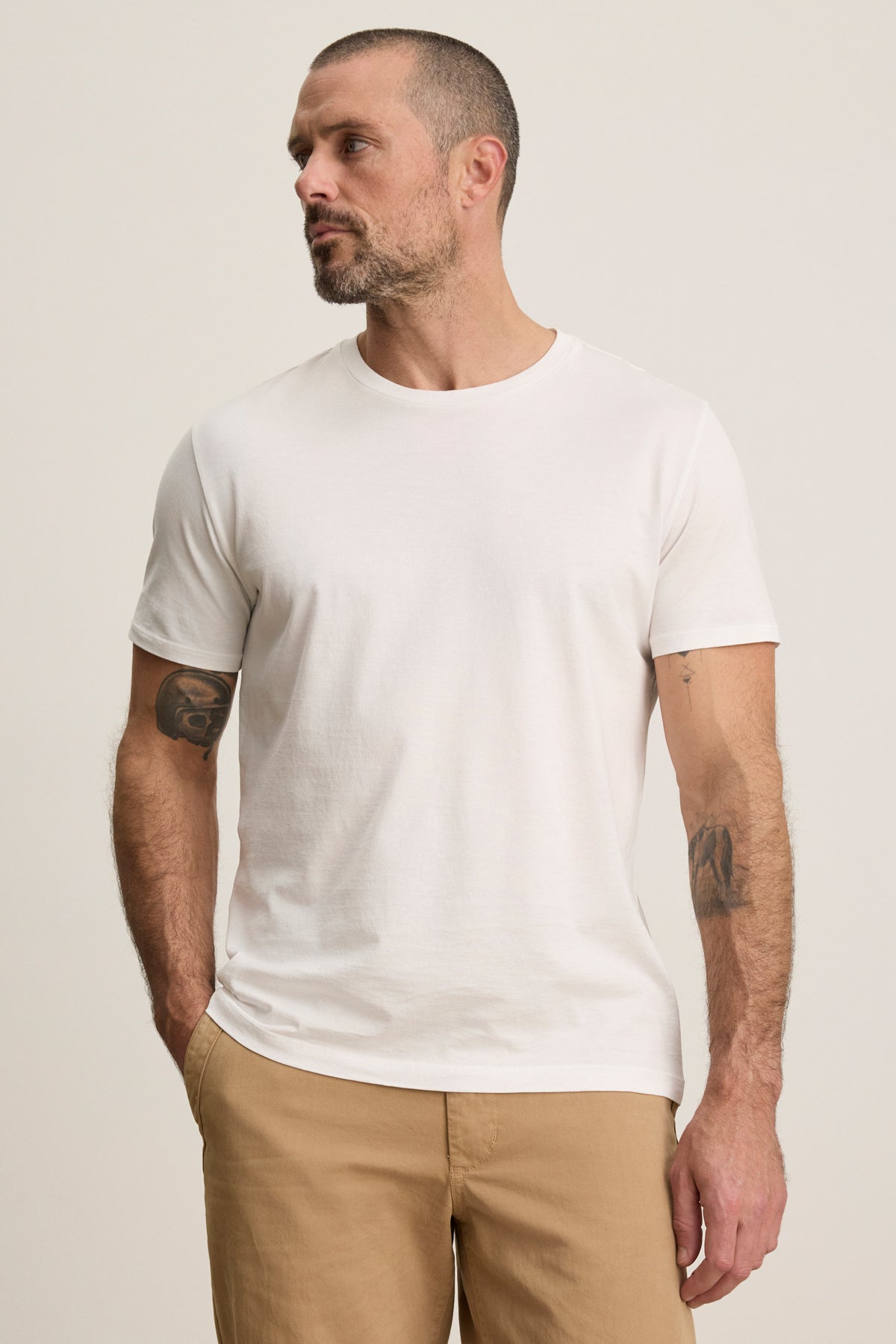 A man wearing the FRANKIE TEE by Velvet by Graham & Spencer—plain white, short-sleeve with tailored sleeves—and beige pants stands with hands in his pockets against a neutral background.-39450521829569