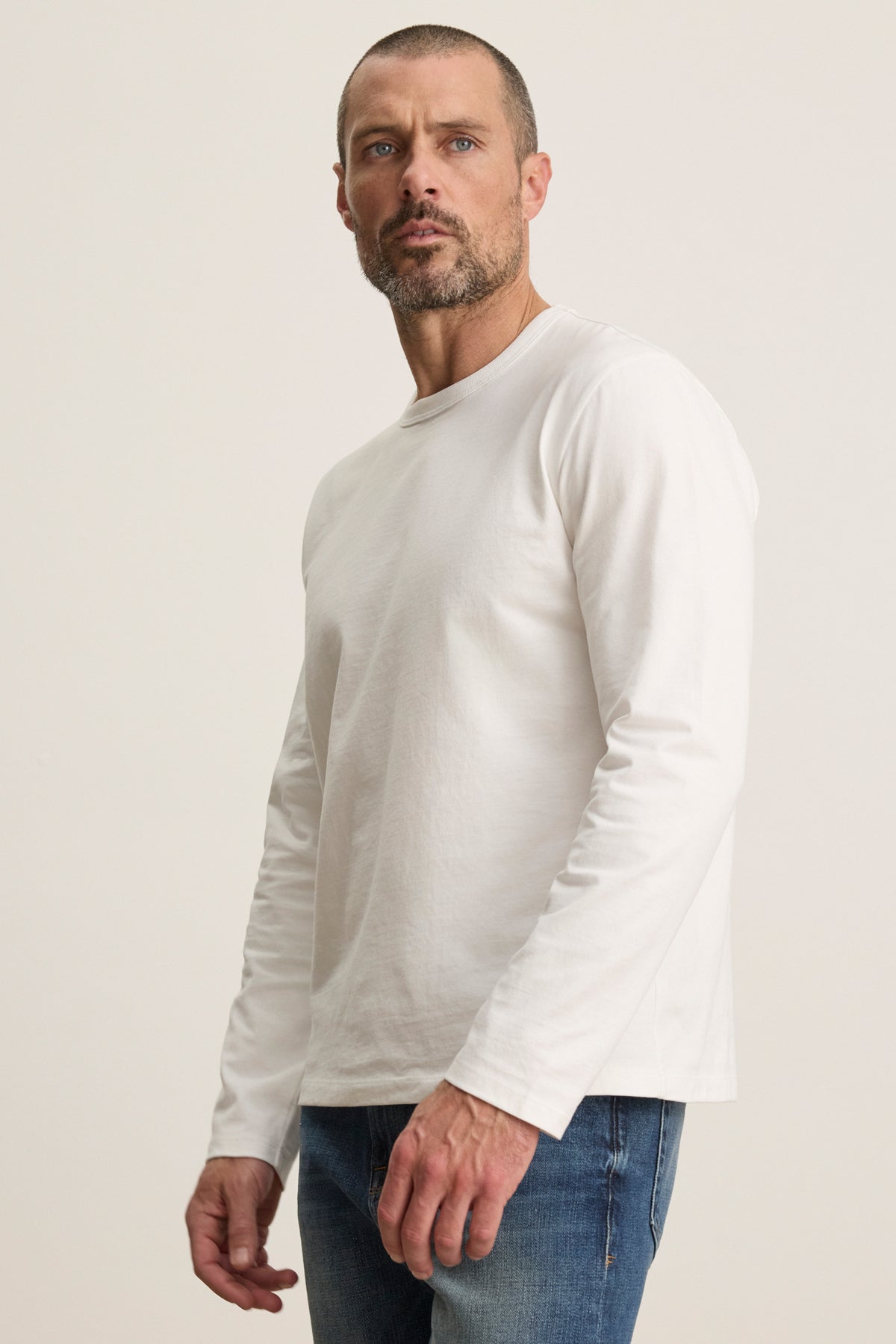 A man with short hair and a beard wears the ANTONIO TEE by Velvet by Graham & Spencer and blue jeans, standing against a neutral background.