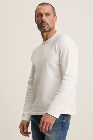 A man with short hair and a beard wears the ANTONIO TEE by Velvet by Graham & Spencer and blue jeans, standing against a neutral background.