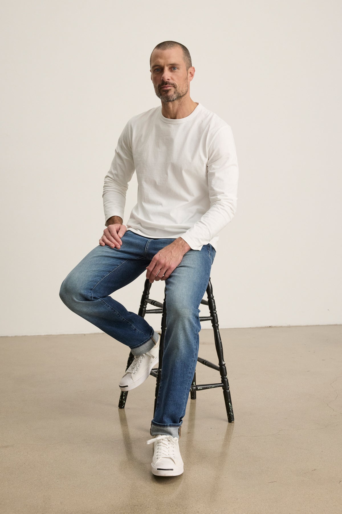 A man with short hair and a beard sits on a black stool, wearing the Velvet by Graham & Spencer ANTONIO TEE, blue jeans, and white sneakers against a plain background.-39543501029569