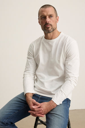 A man with short hair and a beard wears the ANTONIO TEE by Velvet by Graham & Spencer, a relaxed fit white cotton jersey long sleeve, paired with blue jeans as he sits on a chair against a plain background.