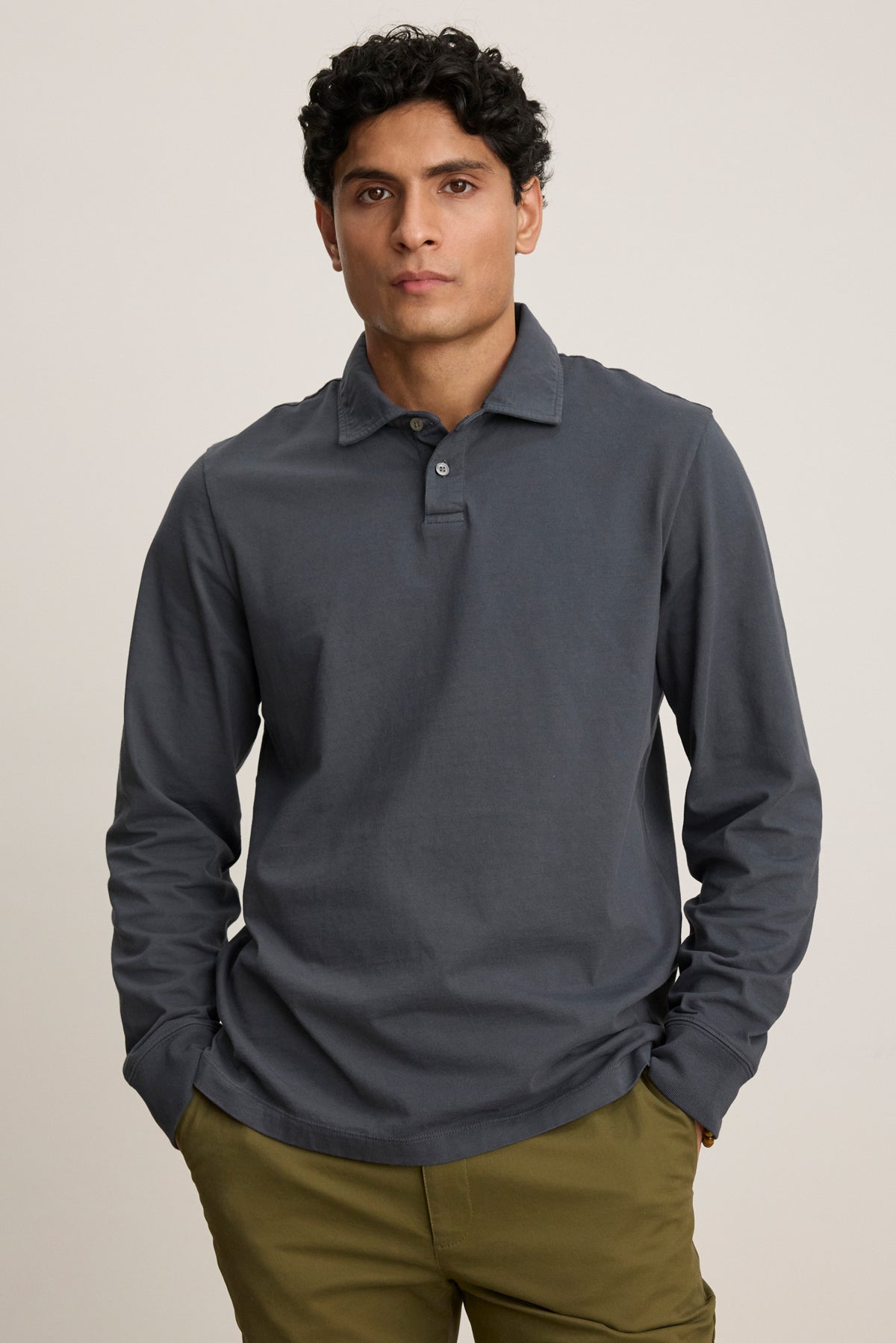 A man stands against a plain background wearing the FLINT POLO by Velvet by Graham & Spencer, a dark gray cotton jersey polo with ribbed cuffs, and olive green pants, his hands in his pockets.-39680471761089