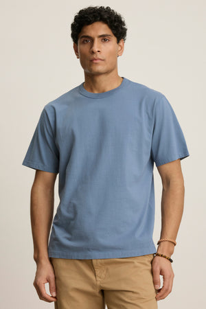 A man with short curly hair wears the Velvet by Graham & Spencer WELLS TEE, a relaxed fit light blue cotton jersey with a ribbed collar, paired with beige pants and standing against a plain light background.