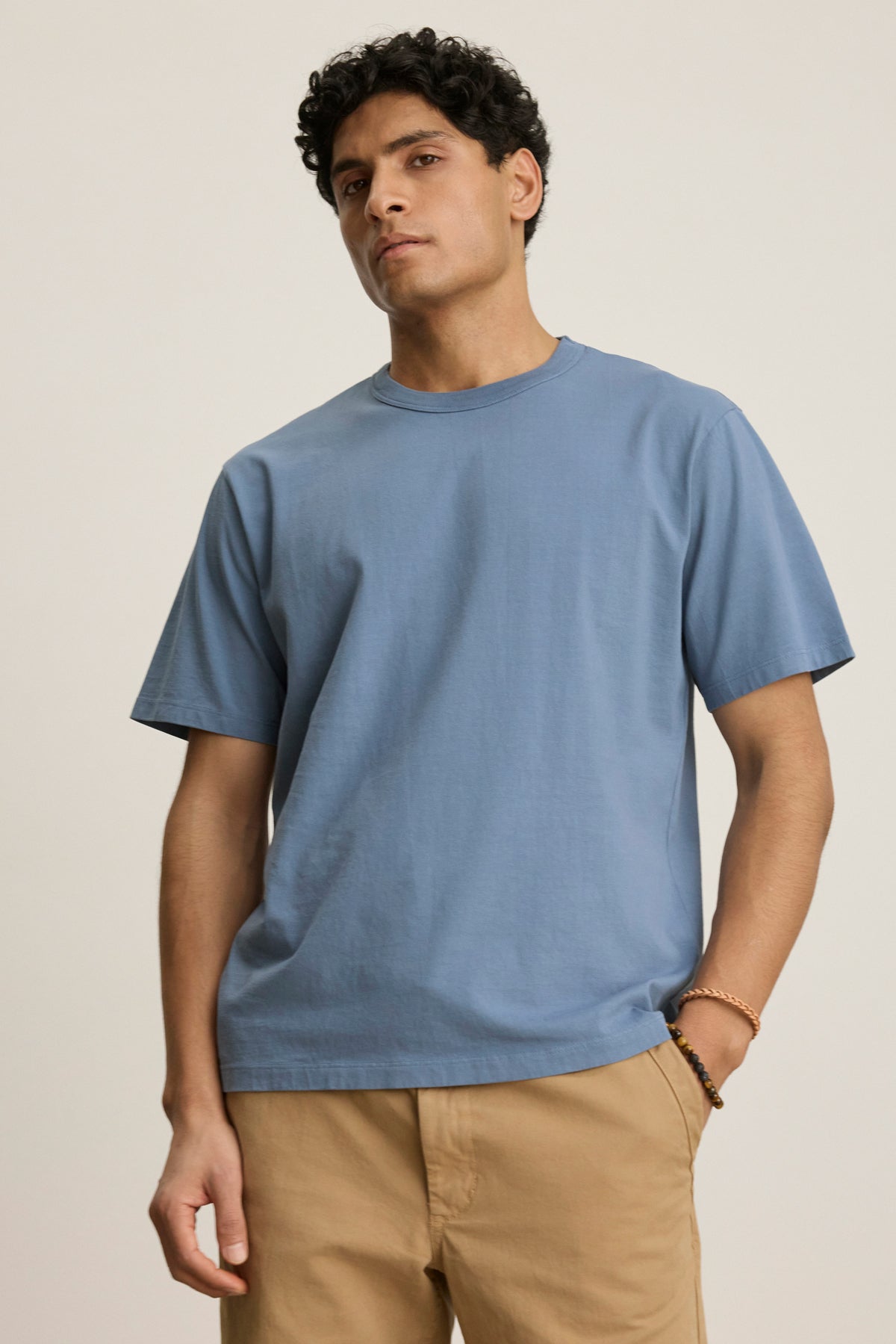 A person stands against a plain background wearing the WELLS TEE by Velvet by Graham & Spencer, a light blue cotton jersey T-shirt with a ribbed collar, paired with beige pants and looking slightly to the side with a neutral expression.-39446052405441