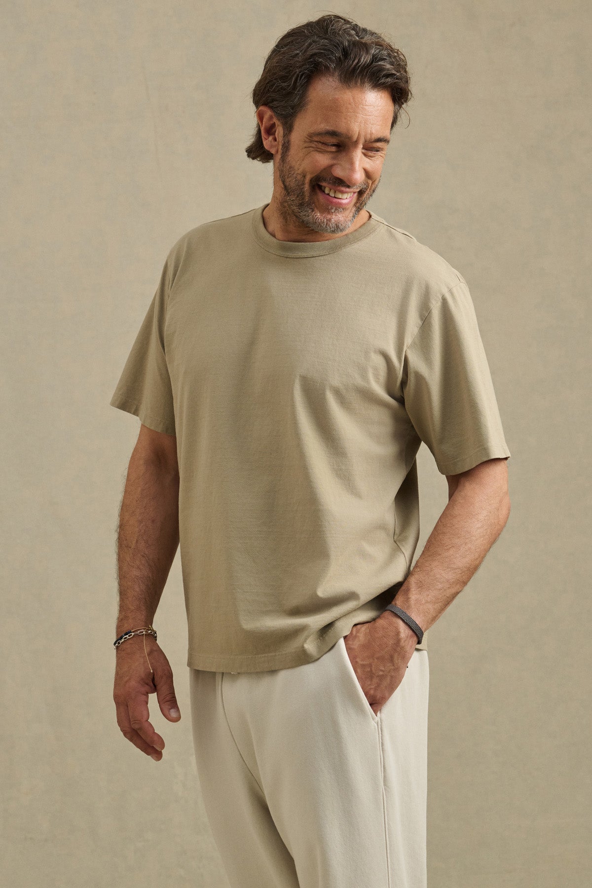 A man with medium-length dark hair and a beard wears the WELLS TEE by Velvet by Graham & Spencer in beige, paired with light pants. He stands smiling with one hand in his pocket—ideal for all-day comfort.-41548224790721