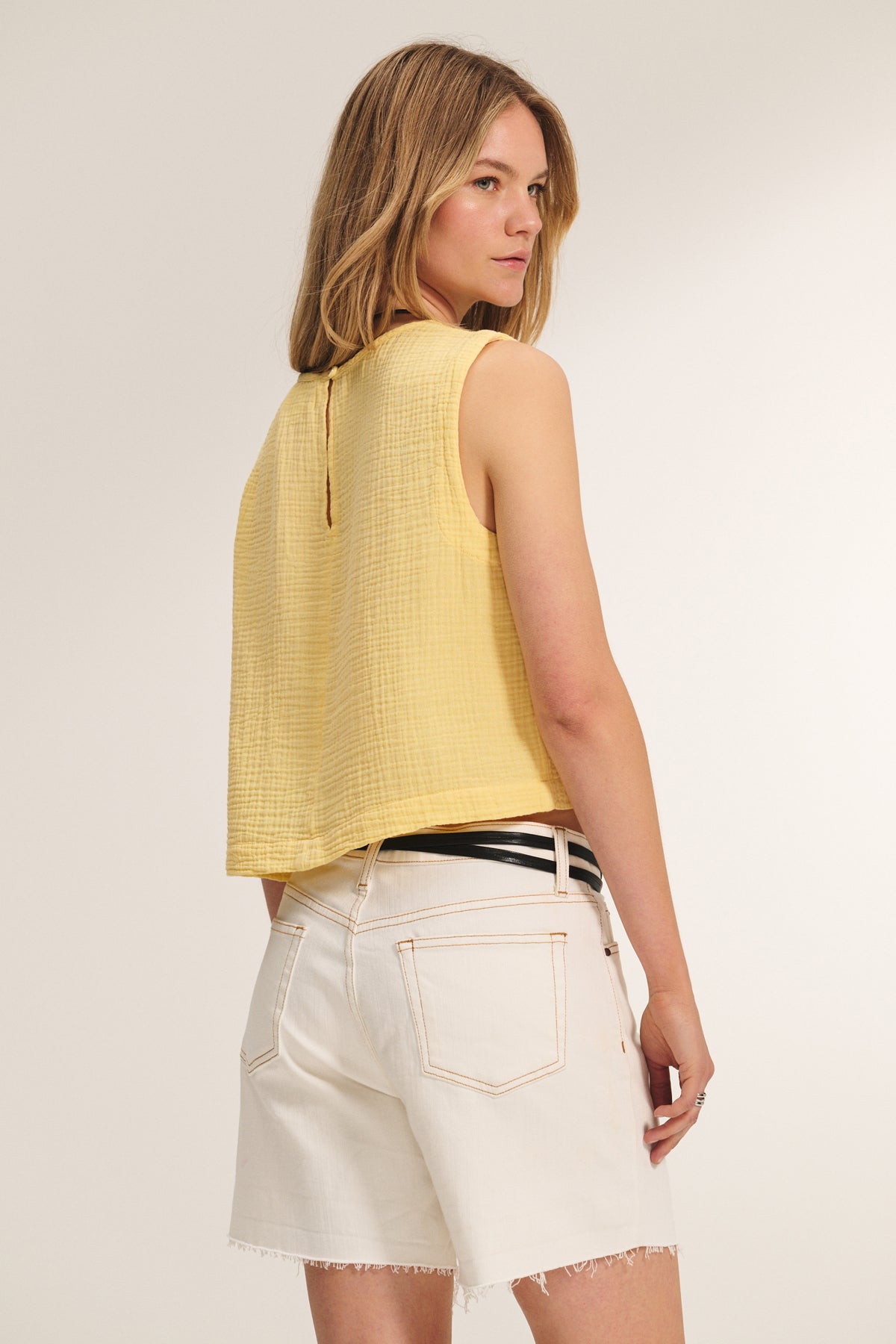 A woman wearing the MILNEY COTTON GAUZE TOP by Velvet by Graham & Spencer and white denim shorts with a black belt stands facing slightly away from the camera.