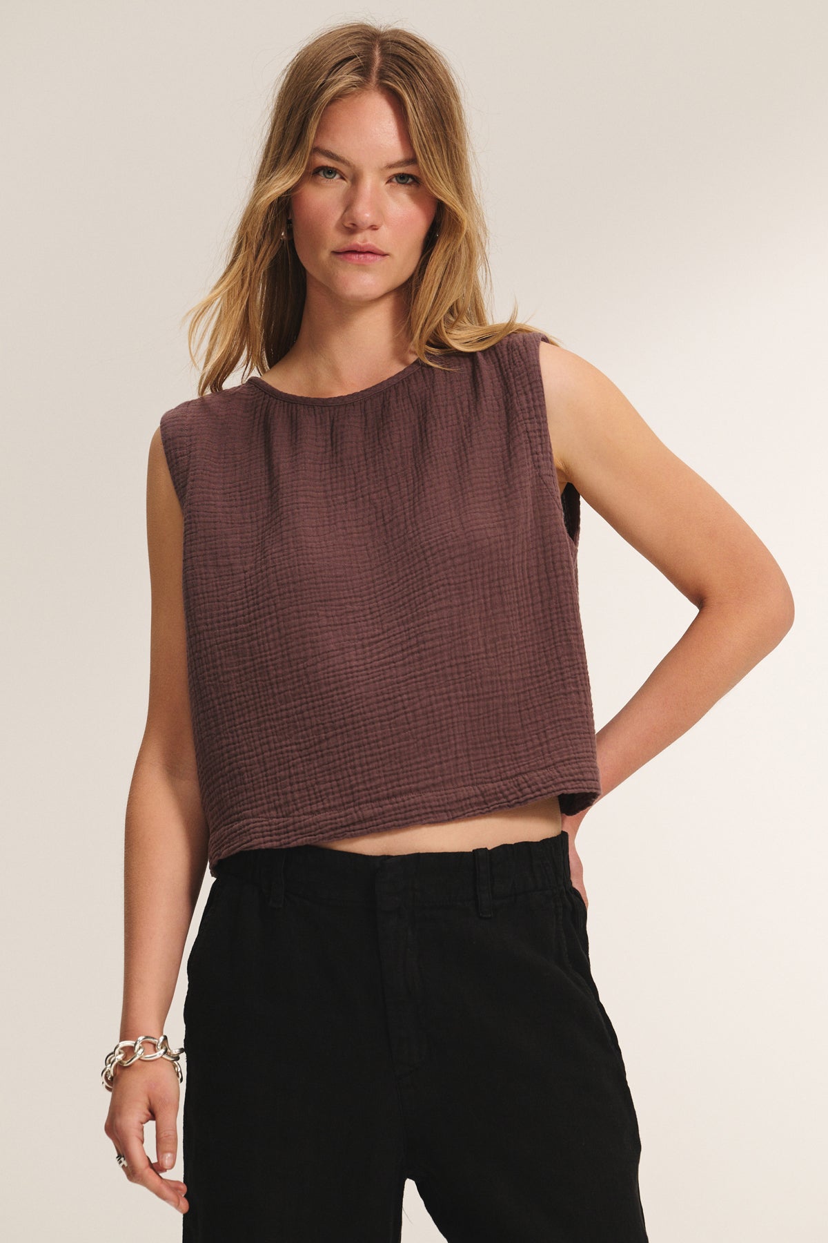 A woman wearing the Velvet by Graham & Spencer MILNEY Cotton Gauze Top, a sleeveless loose-fitting brown textured piece, and black pants stands against a plain background.