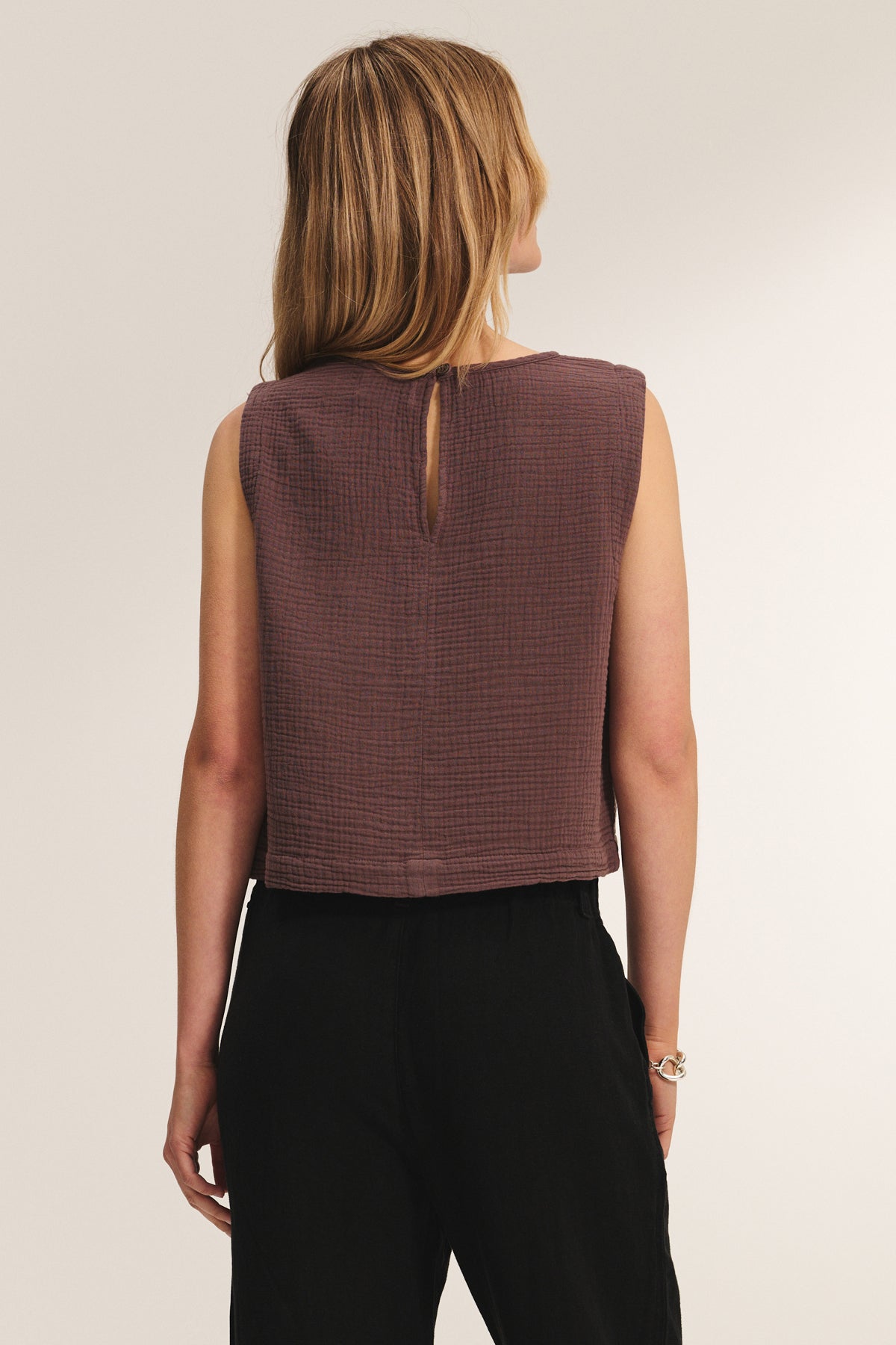 A woman with long, light brown hair is seen from behind wearing the MILNEY COTTON GAUZE TOP by Velvet by Graham & Spencer, a sleeveless brown textured top, paired with black pants against a plain background.-39161095717057