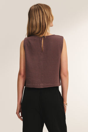 A woman with long, light brown hair is seen from behind wearing the MILNEY COTTON GAUZE TOP by Velvet by Graham & Spencer, a sleeveless brown textured top, paired with black pants against a plain background.