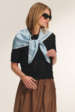 A woman wearing sunglasses, a black short-sleeve knit top, a brown skirt, and the MIRABEL CARDIGAN by Velvet by Graham & Spencer draped over her shoulders.