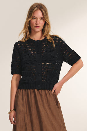 A woman wears the MIRABEL CARDIGAN by Velvet by Graham & Spencer—a lightweight black crochet cardigan with large buttons—paired with a light brown skirt, standing against a neutral background.