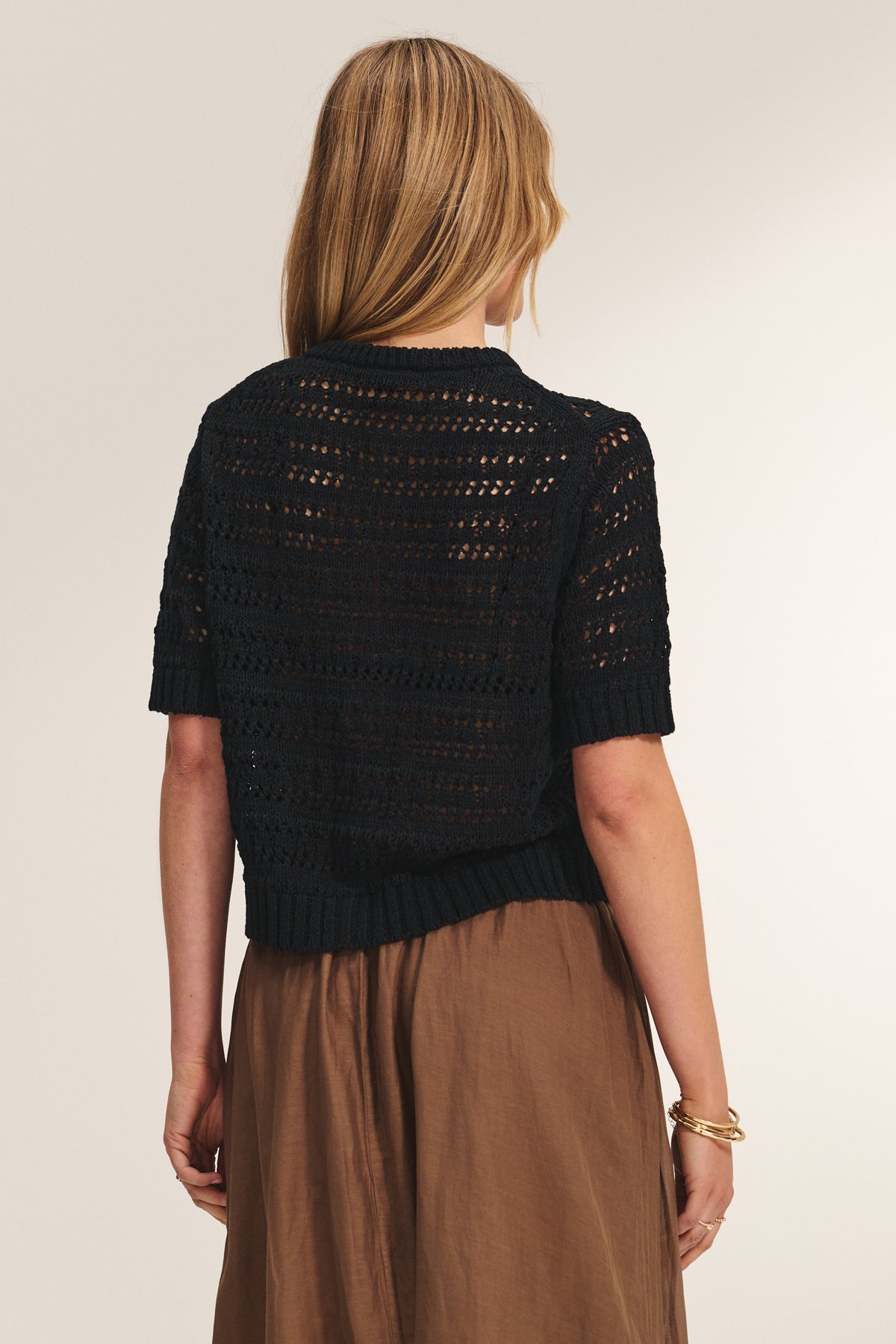 A woman with straight blonde hair wears the MIRABEL CARDIGAN by Velvet by Graham & Spencer, a lightweight black short-sleeve knit sweater, paired with a brown skirt and a gold bracelet on her left wrist.-39149222068417