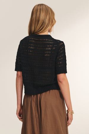 A woman with straight blonde hair wears the MIRABEL CARDIGAN by Velvet by Graham & Spencer, a lightweight black short-sleeve knit sweater, paired with a brown skirt and a gold bracelet on her left wrist.