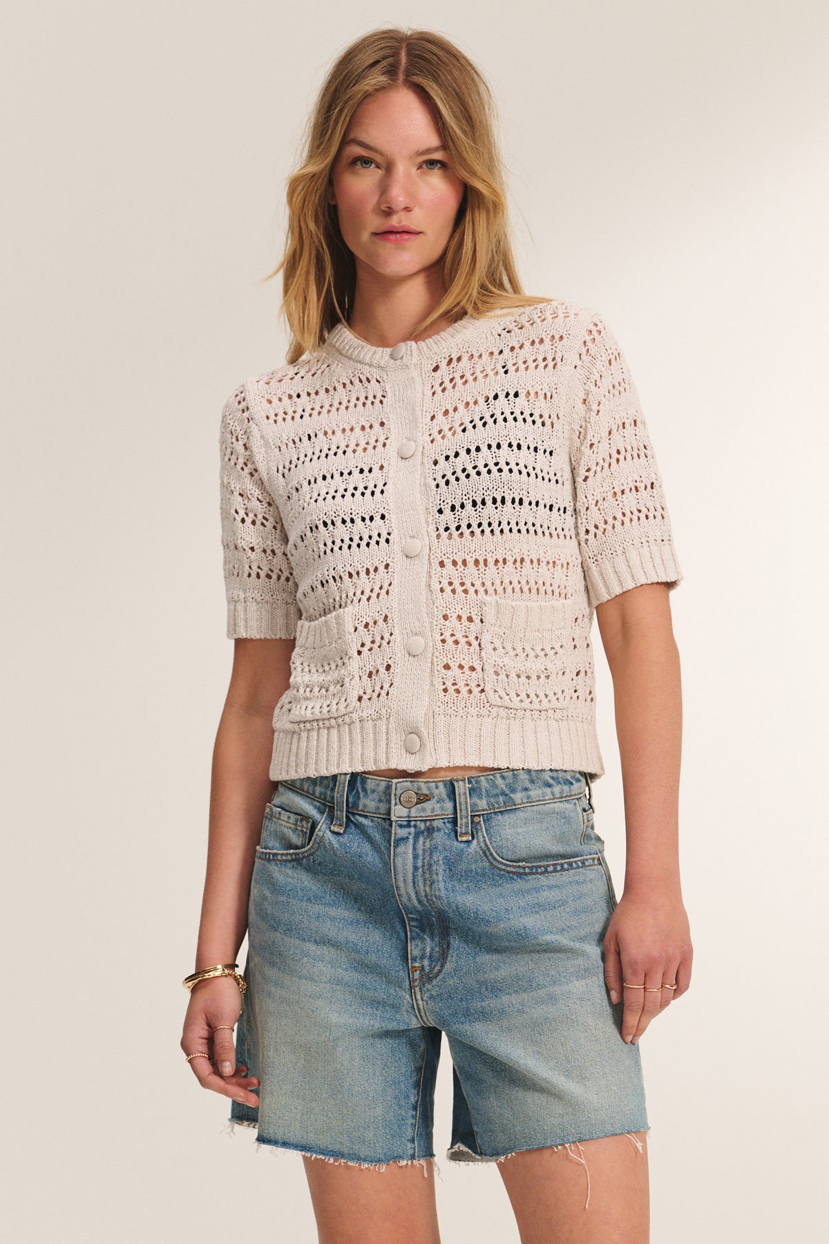 A woman wearing the MIRABEL CARDIGAN by Velvet by Graham & Spencer—a lightweight, beige crochet cardigan with a button-front—paired with blue denim shorts stands against a plain light background.-39149222101185