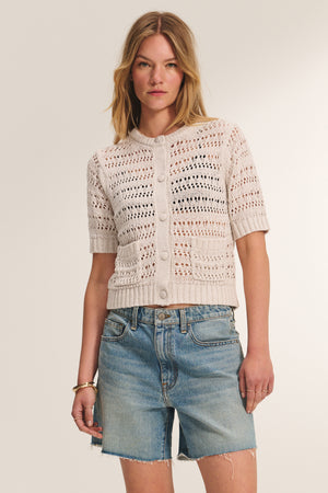 A woman wearing the MIRABEL CARDIGAN by Velvet by Graham & Spencer—a lightweight, beige crochet cardigan with a button-front—paired with blue denim shorts stands against a plain light background.