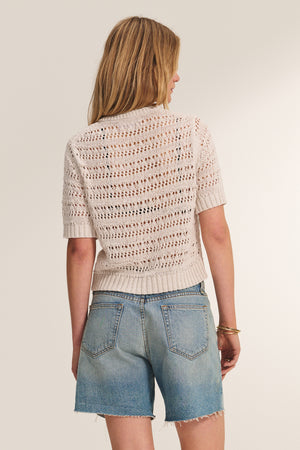 A woman faces away, wearing the MIRABEL CARDIGAN by Velvet by Graham & Spencer in beige crochet with a lightweight feel, paired with light blue denim shorts with frayed hems against a plain background.