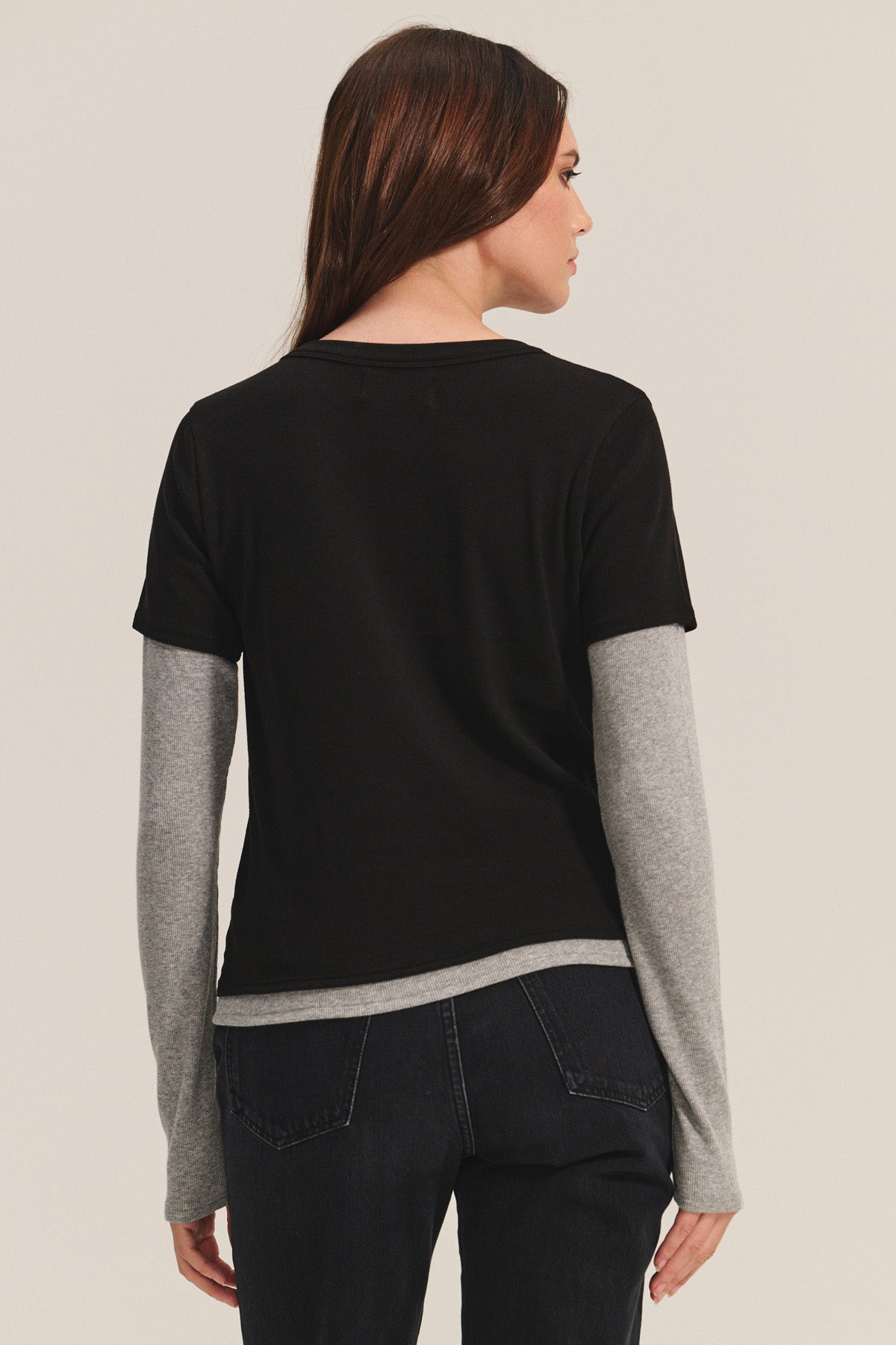 A person with long brown hair is seen from the back, wearing the Velvet by Graham & Spencer BRONSON TEE—a black modal rib crew neck—layered over a gray long-sleeve shirt and paired with dark jeans for a stylish, double-layered look.-40681598615745