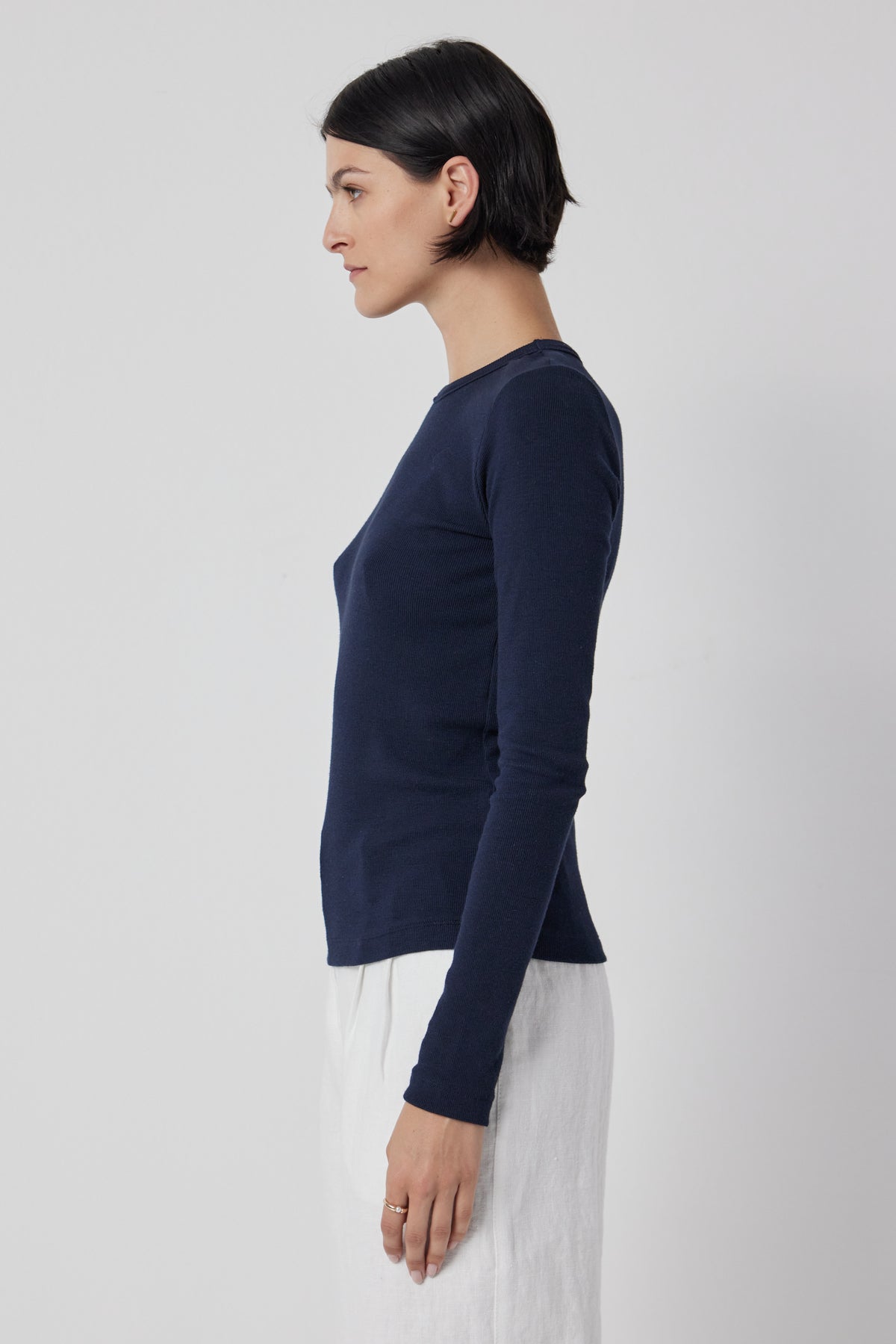 Woman standing in profile wearing a dark blue long-sleeve Camino Tee by Velvet by Jenny Graham and white pants against a neutral background.-36463422013633