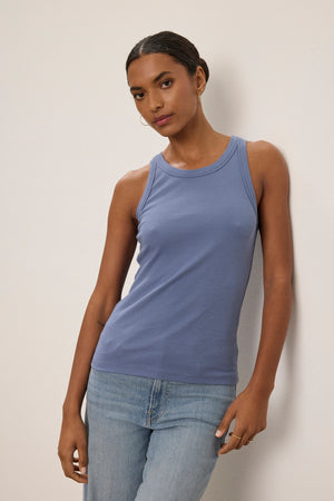 A person wears the CRUZ TANK TOP by Velvet by Jenny Graham and light blue jeans, standing against a plain off-white wall.