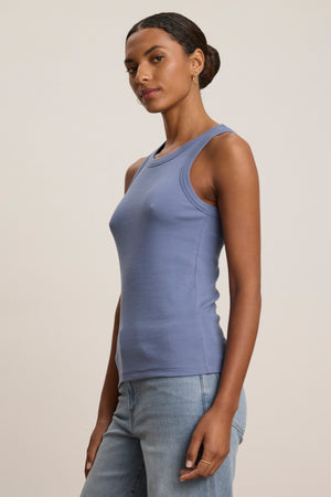 A woman with a neutral expression wears the CRUZ TANK TOP by Velvet by Jenny Graham—a fitted, sleeveless, light blue ribbed cotton-modal blend—paired with light blue jeans against a plain background; a capsule wardrobe essential.