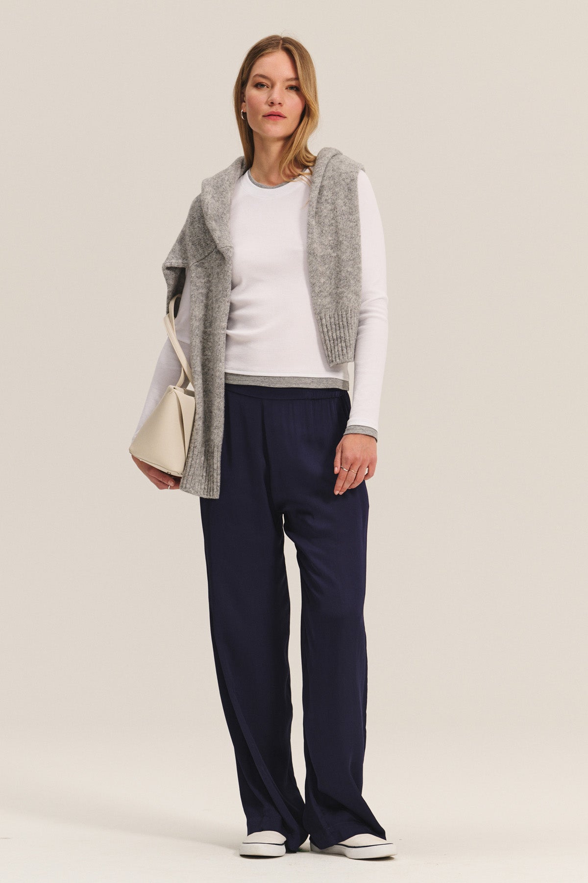 A woman stands facing forward, wearing the NOVATO TEE by Velvet by Graham & Spencer and navy wide-leg pants, with a gray sweater draped over her shoulders. She accessorizes with a light bag and white shoes.-40333255377089