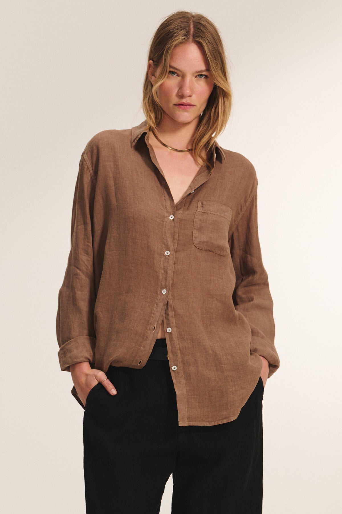 A person wears the MULHOLLAND LINEN SHIRT by Velvet by Jenny Graham—a loose button-up with a chest pocket and relaxed silhouette—paired with black pants, standing hands-in-pockets against a plain background.