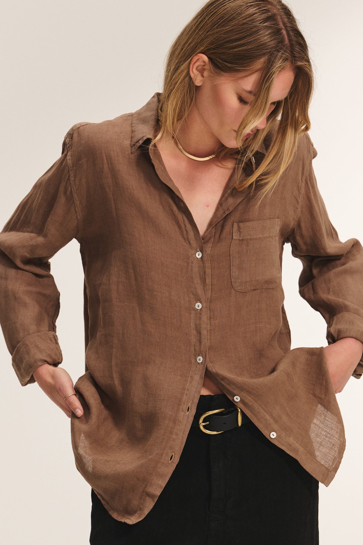 A woman wearing the MULHOLLAND LINEN SHIRT by Velvet by Jenny Graham—a relaxed brown linen button-up with a front pocket—paired with black pants, looks down with her hands in her pockets.