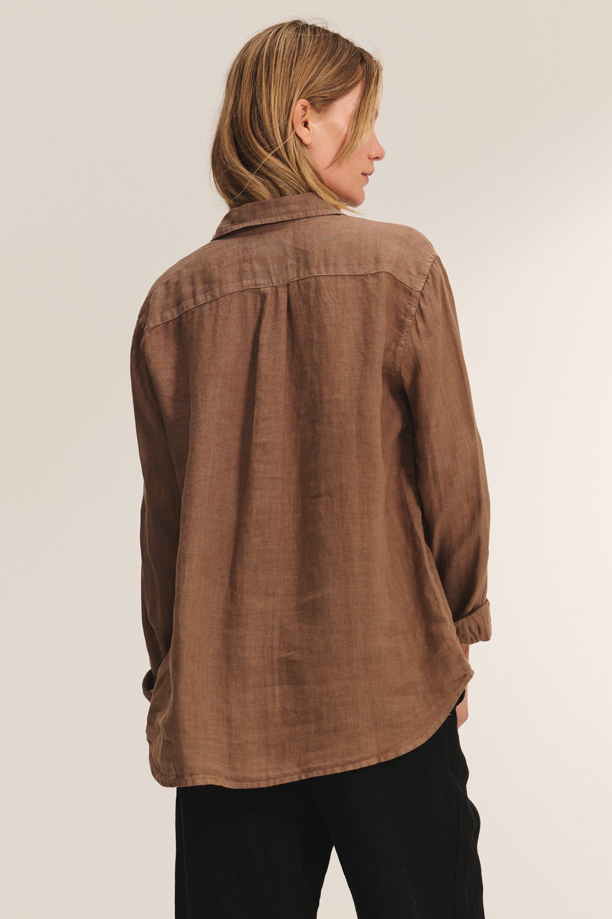 A person with shoulder-length hair is seen from behind wearing the Velvet by Jenny Graham MULHOLLAND LINEN SHIRT in brown and black pants, photographed against a plain background.-39149231046849