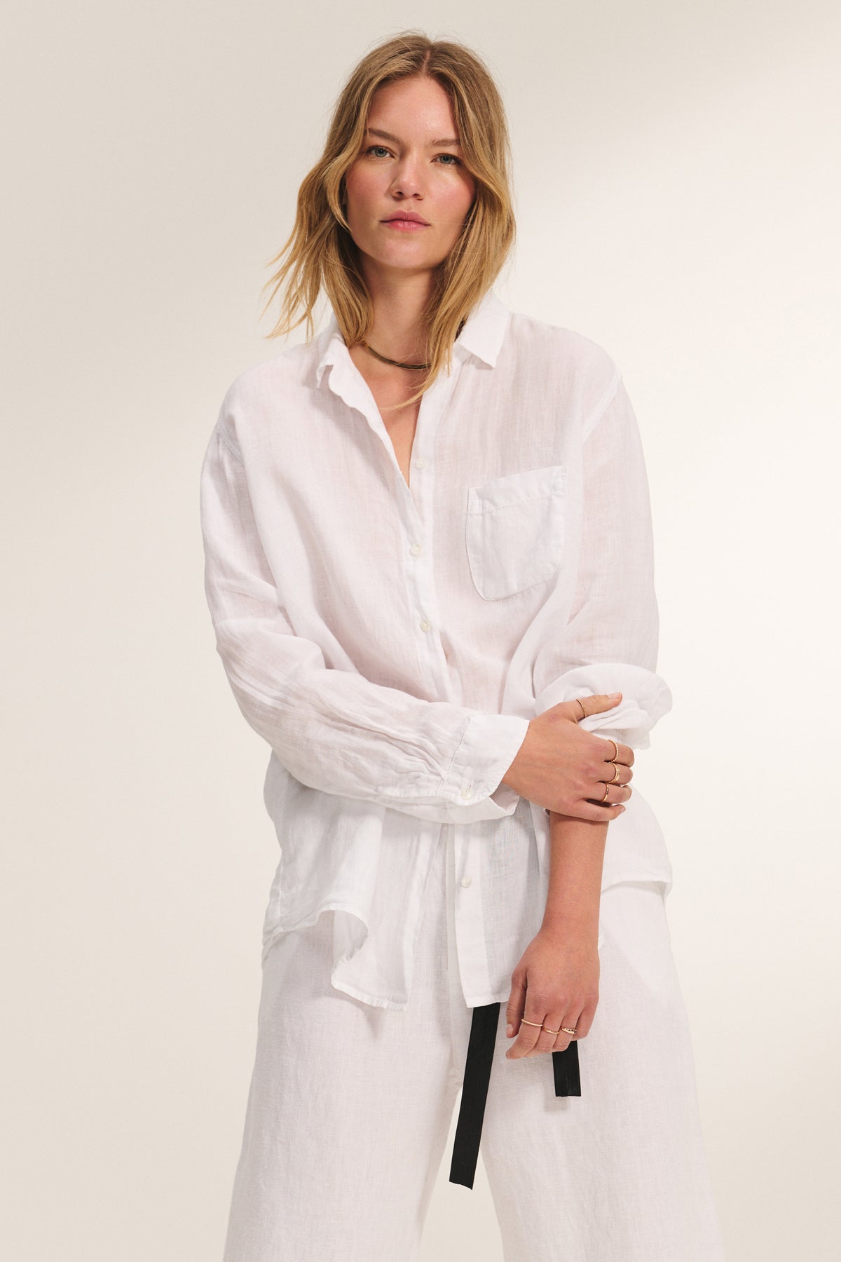 A woman models the MULHOLLAND LINEN SHIRT by Velvet by Jenny Graham—a relaxed, scooped-hem button-up with front pocket—paired with white pants against a plain light background.-39149231112385