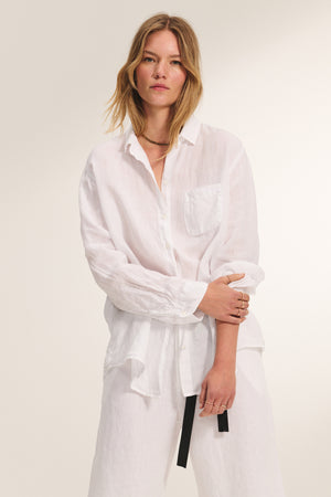A woman models the MULHOLLAND LINEN SHIRT by Velvet by Jenny Graham—a relaxed, scooped-hem button-up with front pocket—paired with white pants against a plain light background.