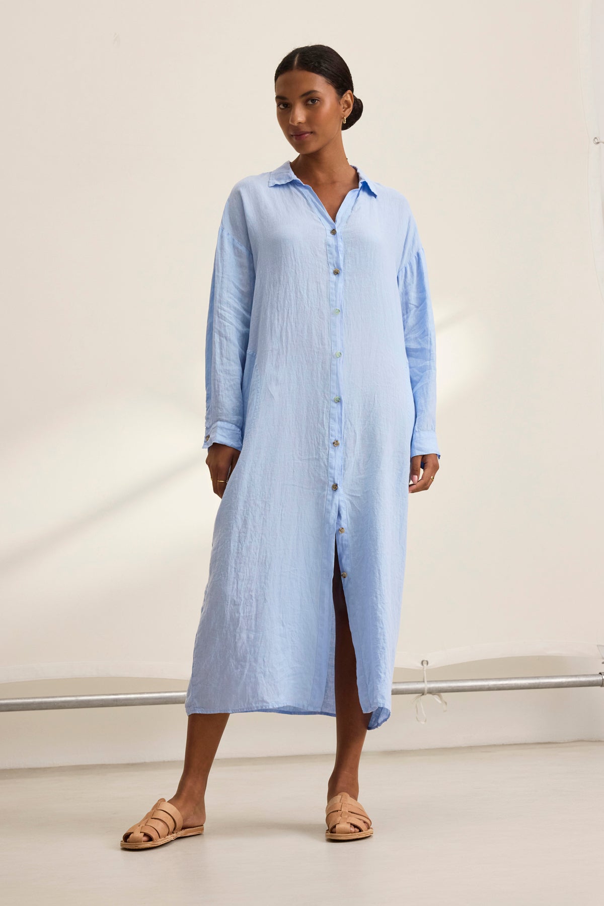 In a neutral indoor setting, someone showcases the breathable comfort and versatile design of the Velvet by Graham & Spencer NICOLE LINEN DRESS in light blue, paired with tan sandals.