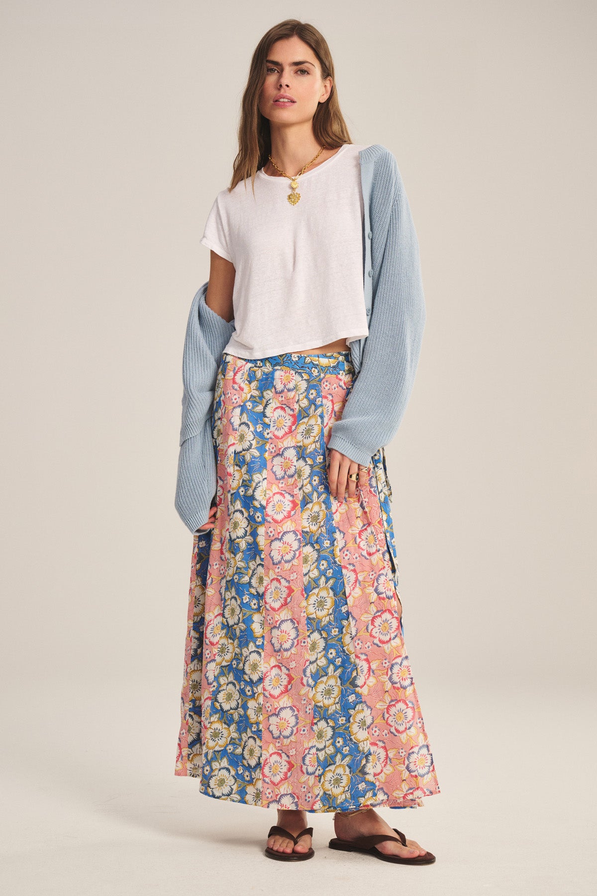 A woman wears the NICOLETTE SKIRT by Velvet by Graham & Spencer—a printed cotton maxi skirt with pink and blue patterns—paired with a white t-shirt, light blue cardigan over her arms, and black sandals.-41008639475905