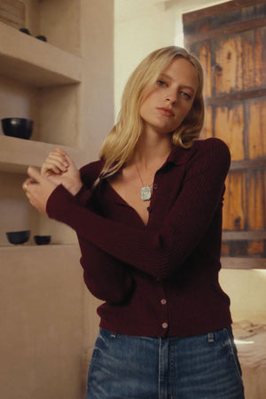 A woman with long blonde hair wears the NIKKI COTTON CASHMERE CARDIGAN by Velvet by Graham & Spencer in burgundy, paired with blue jeans, standing indoors near shelves and a wooden window.