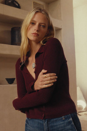 A woman with long blonde hair wears the NIKKI COTTON CASHMERE CARDIGAN by Velvet by Graham & Spencer in burgundy with blue jeans, crossing her arms while standing indoors in front of shelves filled with pottery.