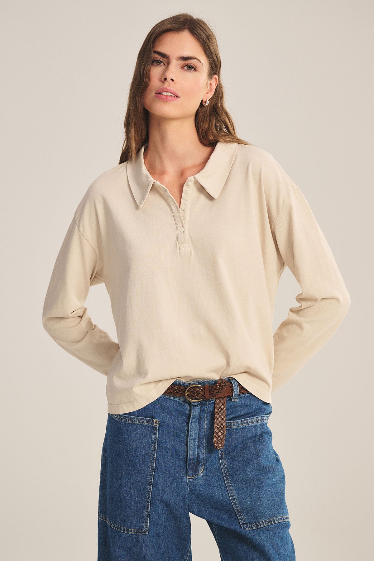 A woman wears the NILLI POLO by Velvet by Graham & Spencer—a vintage cotton jersey, boxy-fit polo with drop shoulders—tucked into high-waisted blue jeans and styled with a brown braided belt against a plain background.-39654977667265