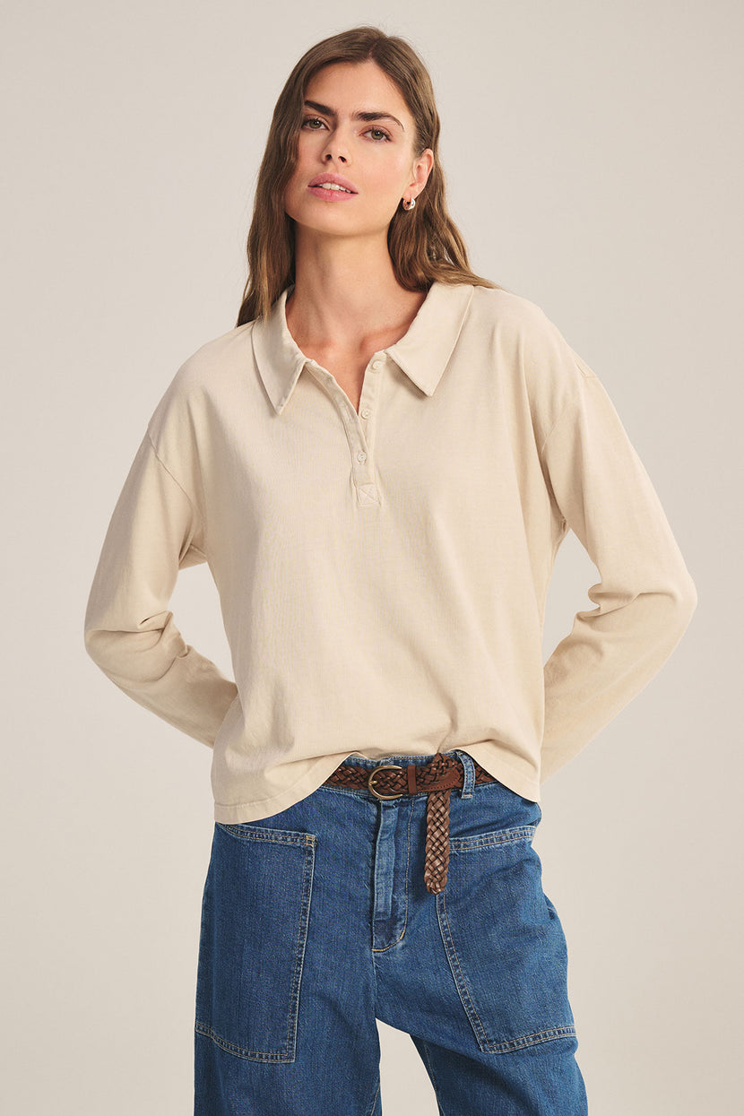 A woman wears the NILLI POLO by Velvet by Graham & Spencer—a vintage cotton jersey, boxy-fit polo with drop shoulders—tucked into high-waisted blue jeans and styled with a brown braided belt against a plain background.