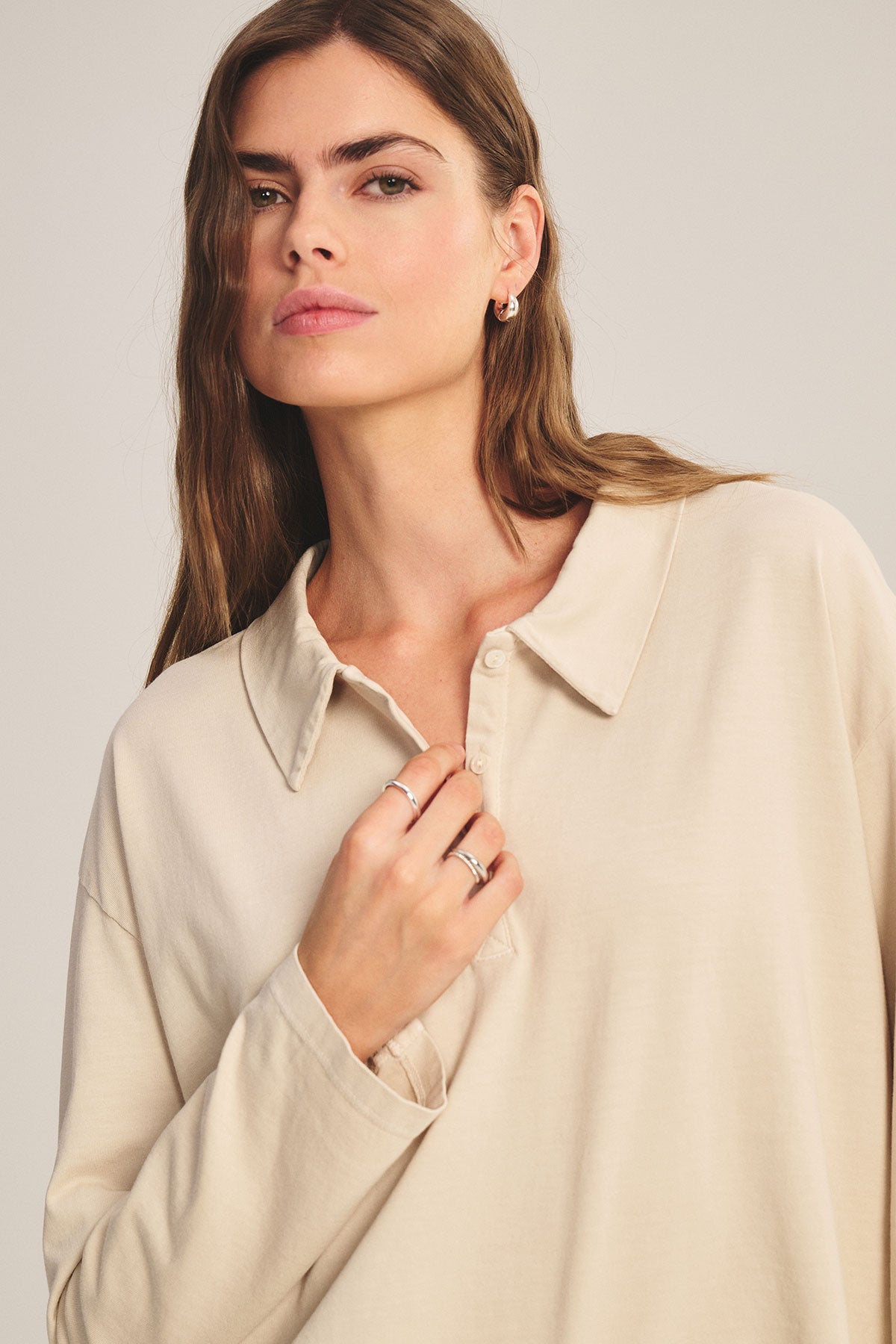 A woman with long brown hair wears the NILLI POLO by Velvet by Graham & Spencer, a beige boxy fit top with drop shoulders and minimalist jewelry, looking slightly to the side with a neutral expression.-39654977700033