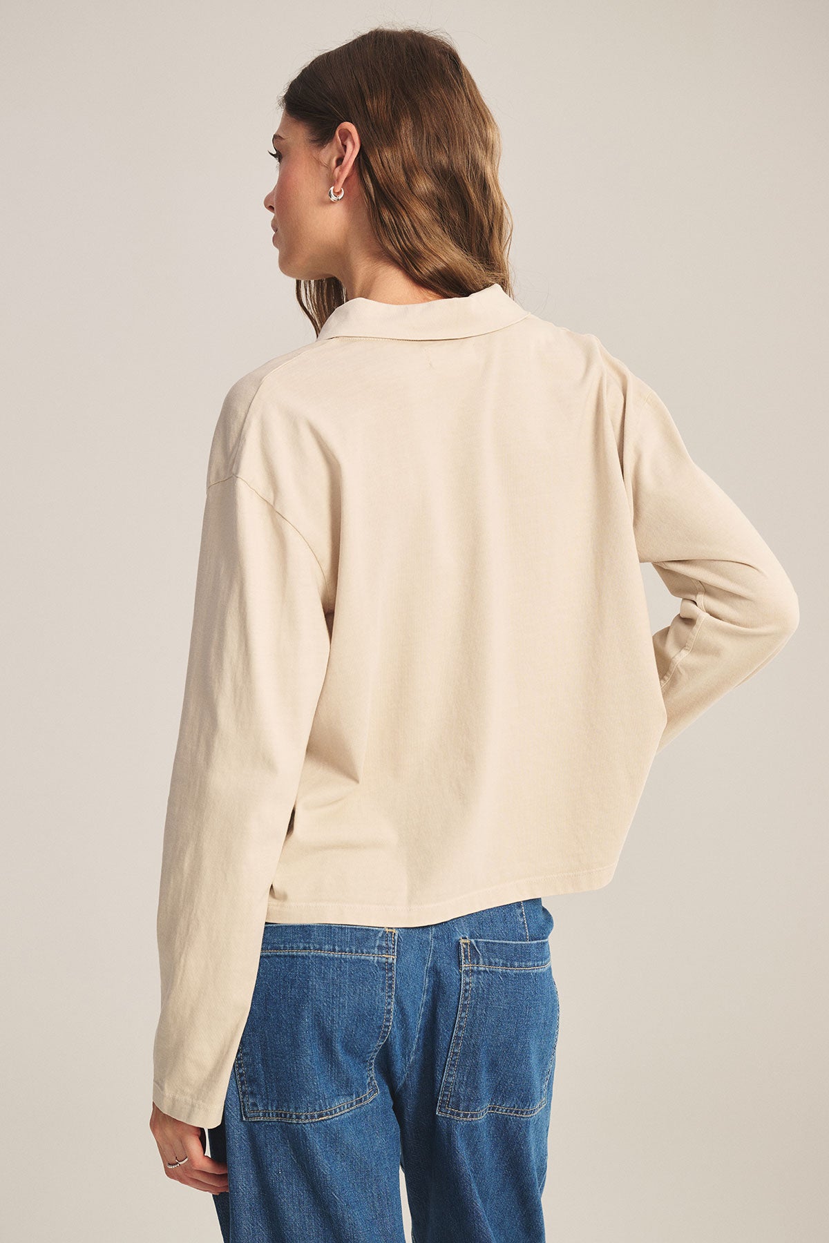 A woman faces away against a neutral background, wearing the NILLI POLO beige vintage cotton jersey long-sleeve shirt by Velvet by Graham & Spencer and blue jeans with visible back pockets.