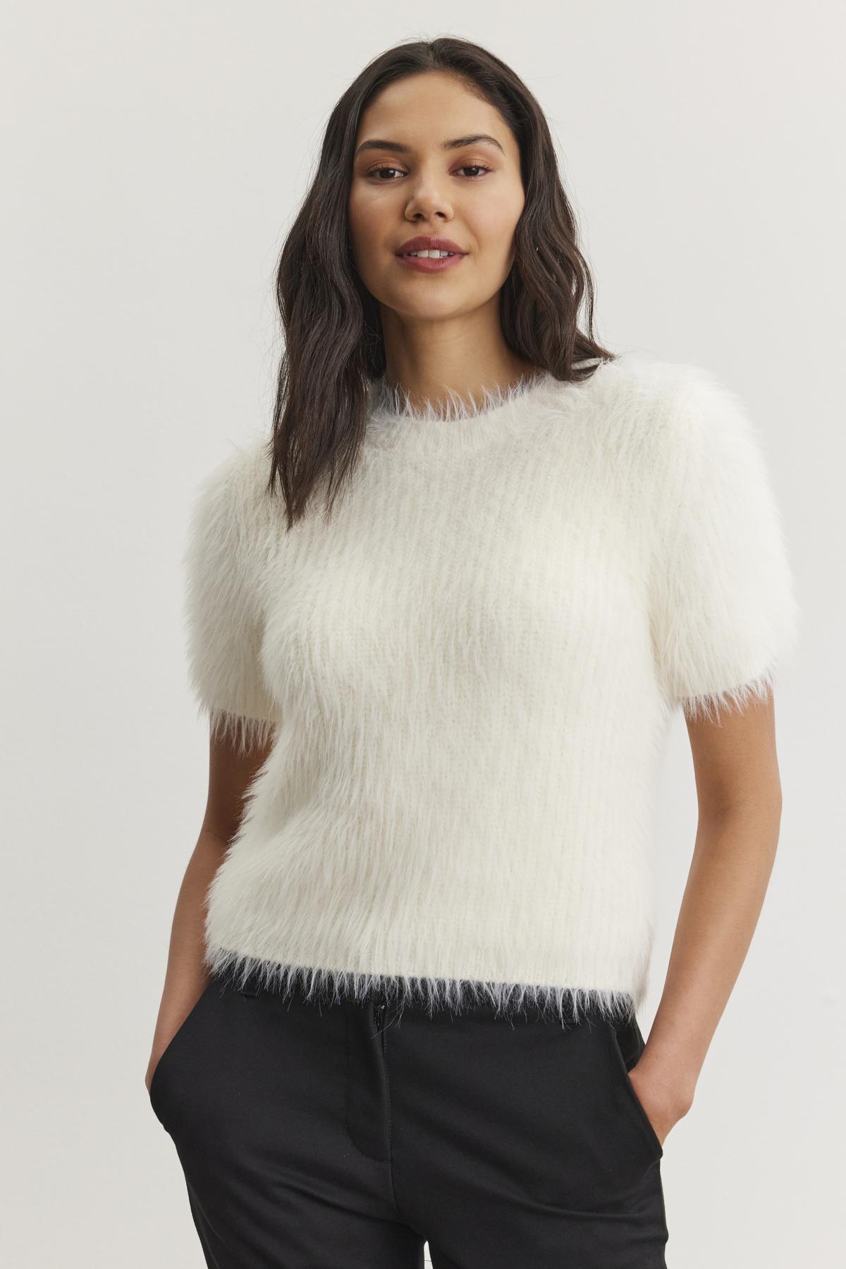A person wearing the EILA FEATHER YARN SWEATER by Velvet by Graham & Spencer, featuring a white, short-sleeved design with a fuzzy texture, pairs it with black pants and stands with hands in pockets against a plain background.-38246411567297