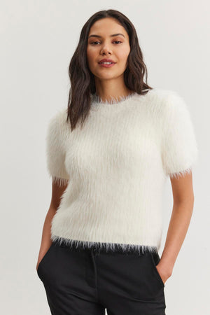 A person wearing the EILA FEATHER YARN SWEATER by Velvet by Graham & Spencer, featuring a white, short-sleeved design with a fuzzy texture, pairs it with black pants and stands with hands in pockets against a plain background.