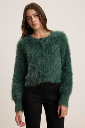 A person wearing a Holland Feather Yarn Cardigan by Velvet by Graham & Spencer and black pants stands against a plain backdrop, showcasing their versatile style.