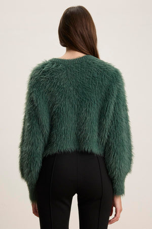 A person with long brown hair is wearing a green HOLLAND FEATHER YARN CARDIGAN by Velvet by Graham & Spencer and black pants, viewed from the back against a plain background.