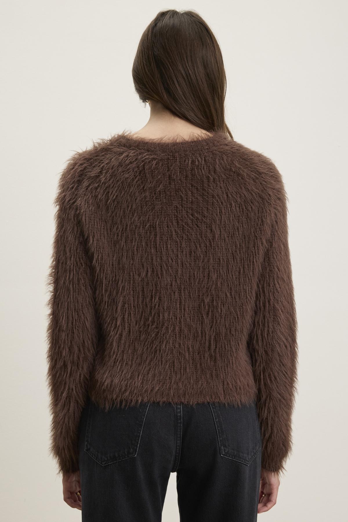 A person is seen from behind wearing the MAYLIE FEATHER YARN SWEATER from Velvet by Graham & Spencer, characterized by its brown feather yarn material and crew neckline, paired with dark jeans against a light background.