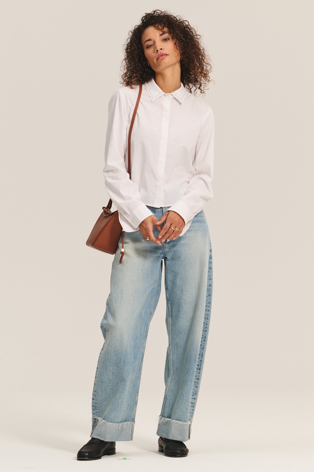 A person stands against a plain background wearing the JOCELYN SHIRT by Velvet by Graham & Spencer, paired with light blue wide-leg jeans with cuffs, black shoes, and carrying a brown shoulder bag.-40247705567425