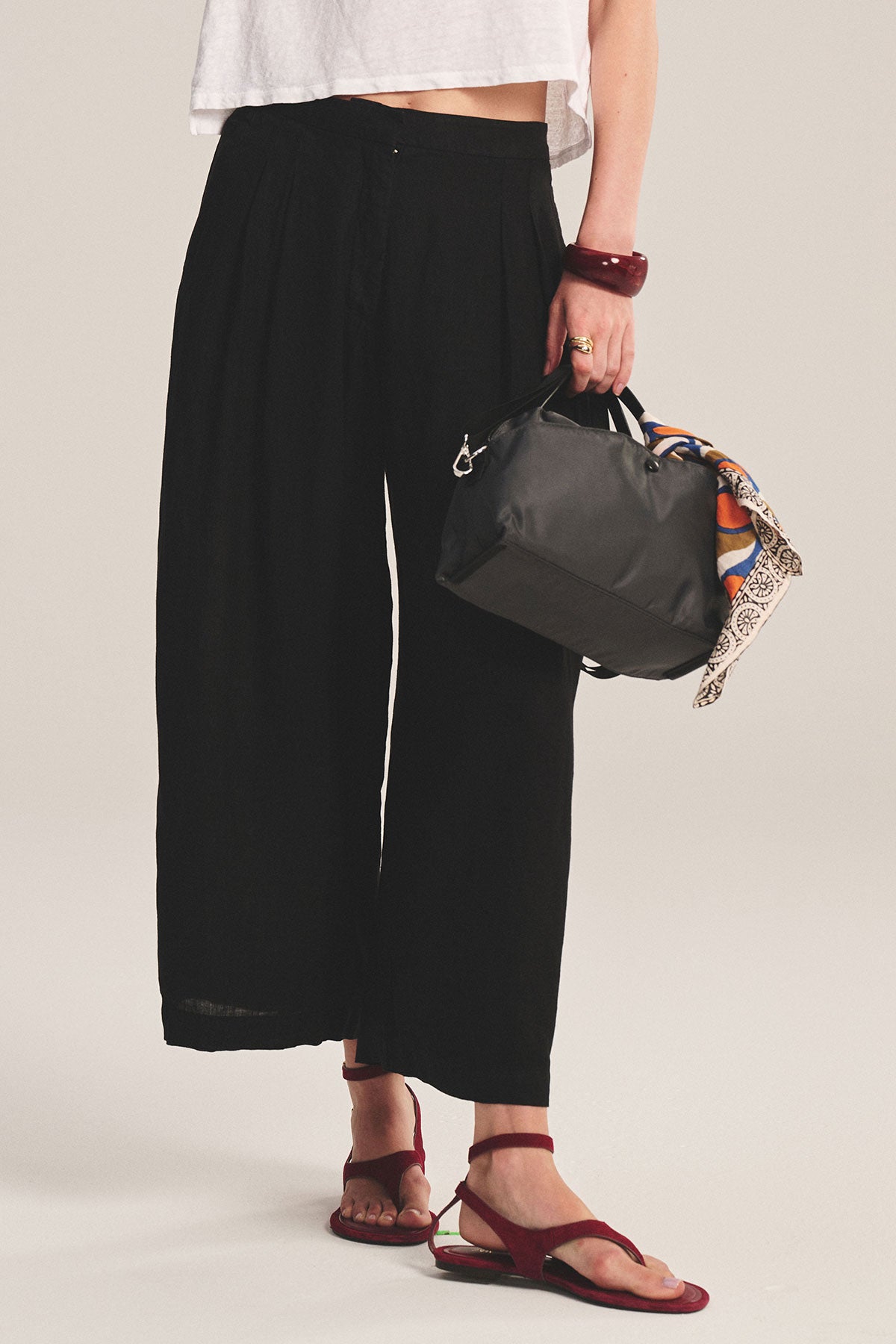A person wears a white crop top, black wide-leg pants, and red sandals while holding the Velvet by Graham & Spencer NYLON CROSSBODY BAG with a patterned scarf tied on—ideal for everyday use.