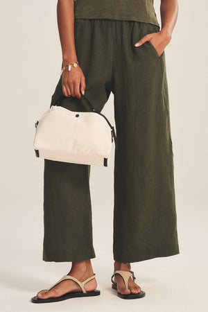 Wearing olive green wide-leg pants and sandals, they carry the Velvet by Graham & Spencer NYLON CROSSBODY BAG—a small beige everyday essential—with one hand in their pocket.