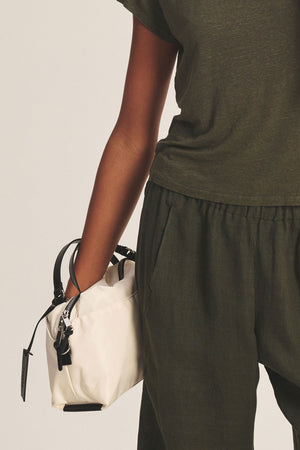A person in an olive green outfit holds the Velvet by Graham & Spencer NYLON CROSSBODY BAG— a small white bag with black handles, shown by their side against a plain background, ideal for everyday use.