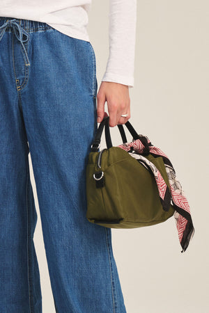 A person in blue jeans and a white top carries the NYLON CROSSBODY BAG by Velvet by Graham & Spencer—an olive-green bag with black handles and a patterned scarf, ideal for everyday use.